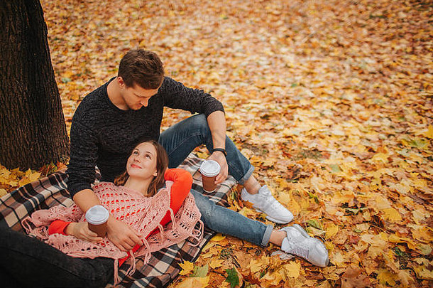 这对年轻夫妇在公园里的可爱照片。躺在毯子上的女人看着那个家伙。男人坐着看着女人。他们有几杯咖啡。