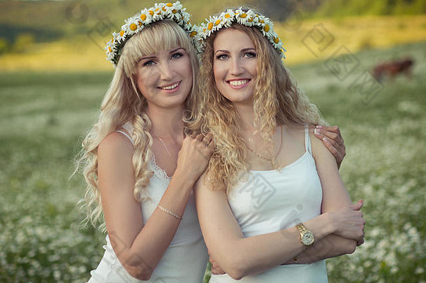 两位可爱的金发少女，一位乳白色皮肤的女士，头戴一个低矮的花环，在台上的草地上享受着一起的时光