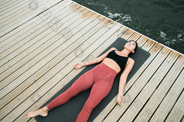 在湖边的码头上冥想的年轻女孩。她穿着黑色上衣和红色紧身裤。