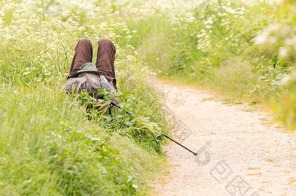 夏天，在一个郊野公园里，脸上戴着帽子，一边拿着一根登山杖，坐在长凳上休息的人
