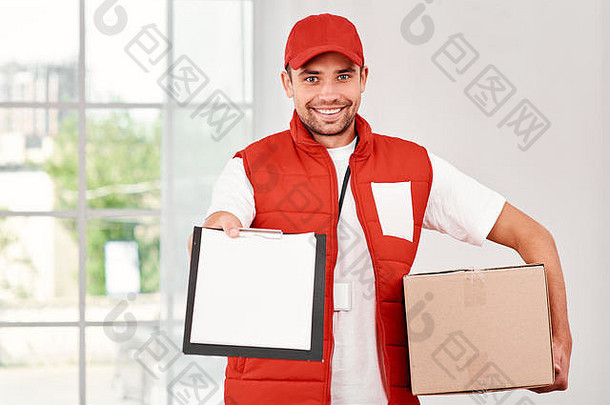 穿着红色邮政制服的快活的邮递员正在给客户递送包裹。他拿着纸箱，等<strong>待发货</strong>时的签名，微笑着看着相机。友好的员工，优质的送货服务。在室内明亮的内部。