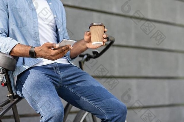 无法辨认的黑人坐在自行车上，用智能手机和咖啡放松