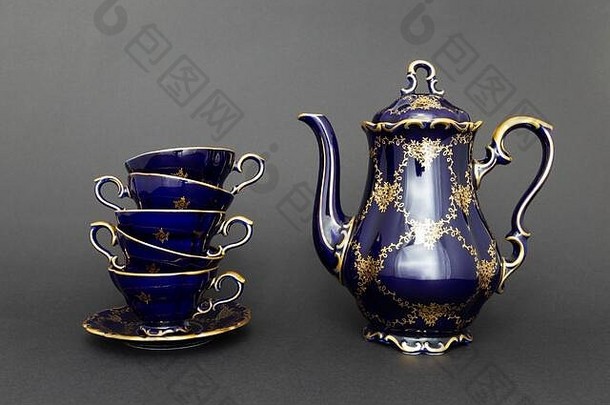 一套美丽的钴蓝色复古瓷茶具的特写镜头，深灰色背景上有金色的花朵图案。这套包括一个茶壶，一个茶壶