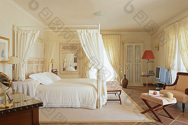 西班牙卧室简单四柱床上的白色窗帘和床单，瓷砖地板上的奶油地毯
