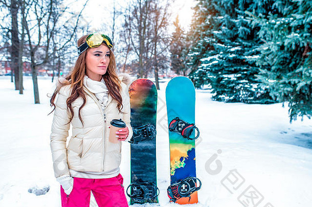 美丽的冬季女子公园，长发保暖的衣服，拿着装有热饮料的玻璃杯，滑雪板，免费的文字开放空间。热咖啡茶