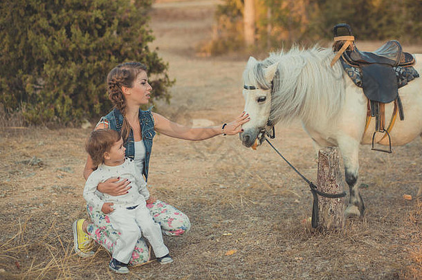 浪漫的场景美丽的女士年轻的母亲和她可爱的小女儿一起在城中村公园散步喂养白马小马。