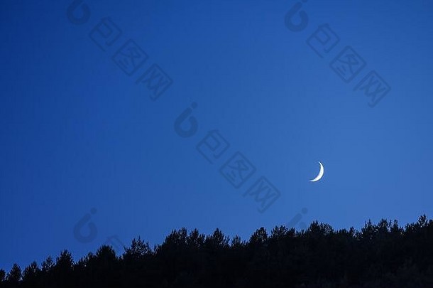 薄薄的月牙儿在夜幕降临前的蓝天下，它旁边的山林剪影