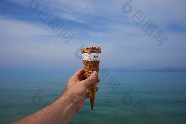 在晴朗的<strong>夏日</strong>，一只手拿着圆锥形的融化的<strong>冰淇淋冰淇淋</strong>，背景是蓝天和湖泊