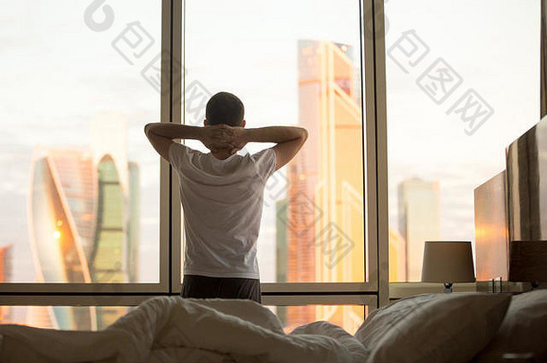 年轻人醒来后在窗口观看黎明城市景色的后视图。早上放松的帅哥