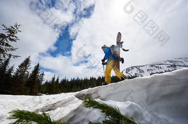 后视图：穿着明亮滑雪服的滑雪运动员背着滑雪板，沿着雪坡爬上山顶。山中探险。背景是蓝天上的云。低角度视图