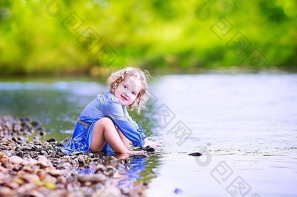 在一个炎热的夏天，穿着蓝色连衣裙的可爱的卷发蹒跚学步的小女孩在河边玩耍，向水中<strong>扔石头</strong>