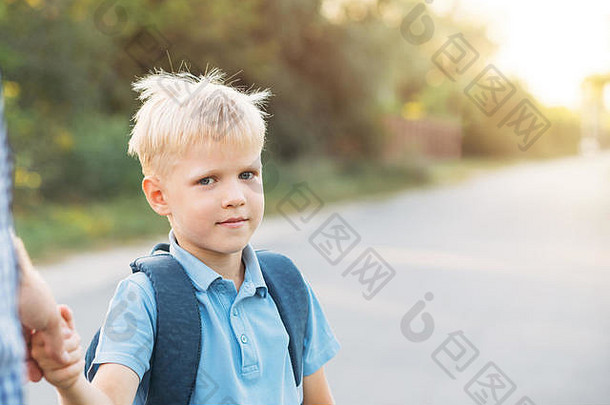 父亲牵着小儿子的手。背着书包的小男孩看着摄像机，正准备上学。回到学校的概念。