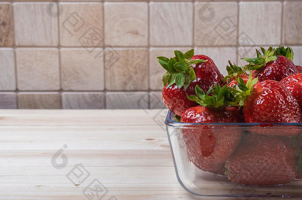 在一张轻薄的木桌上放着一个方形的玻璃碗和草莓。大草莓。