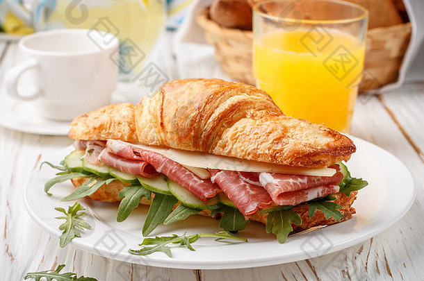 桌上放着新鲜的三明治牛角面包、火腿、芝麻菜、黄瓜和奶酪，还有柠檬水、橙汁和咖啡杯。早餐塞莱茨
