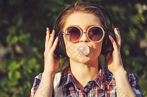 在夏季公园里，年轻的时髦女孩戴着耳机听音乐。嚼口香糖的肖像特写镜头。暖色调。