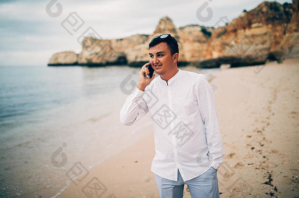 戴眼镜的英俊年轻男子在海滩上散步，用手机聊天。传播观念