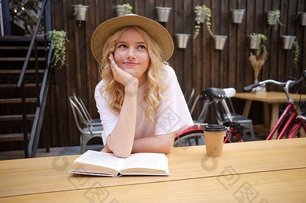 在咖啡厅的庭院里，戴着帽子的美丽浪漫的金发女孩倚在手上梦想着书和咖啡