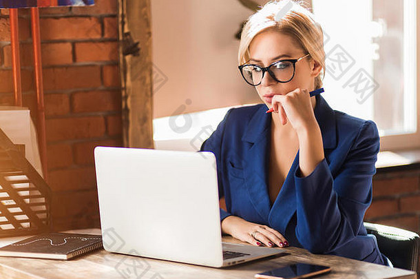 一位年轻漂亮的金发女商人的特写照片，戴着眼镜，穿着时髦的蓝色夹克，在咖啡馆里用白色笔记本电脑工作，手里拿着笔，<strong>严肃认真</strong>