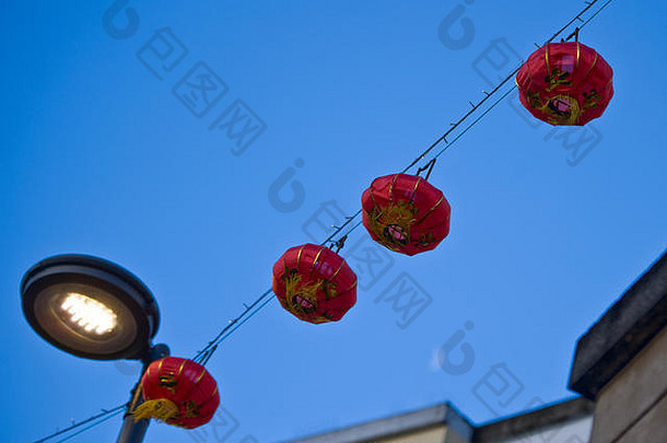 四个红色的中国灯笼挂在电线上，横跨灯柱，在蓝色天空的映衬下，月亮在右下角略微可见。
