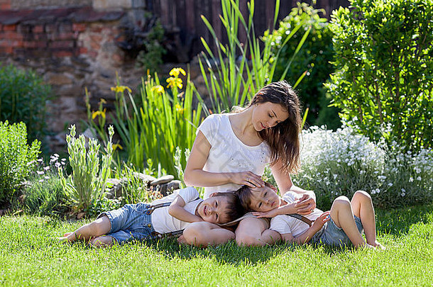 年轻的母亲和她的两个孩子，男孩，坐在草坪上，温柔。妈妈抱着他们