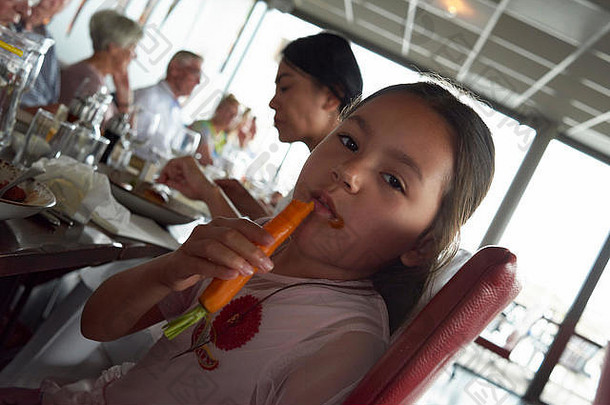 在一家繁忙的餐馆里，一位可爱的年轻亚洲女孩坐在桌子旁，享受着新鲜健康的胡萝卜