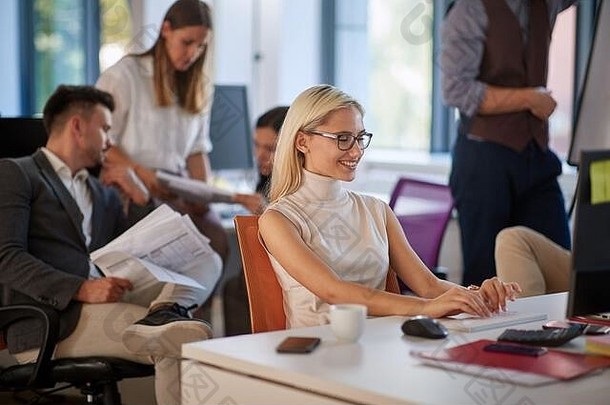 年轻的金发白种人在现代办公室里用电脑打字，背景是一群同事。休闲现代商务