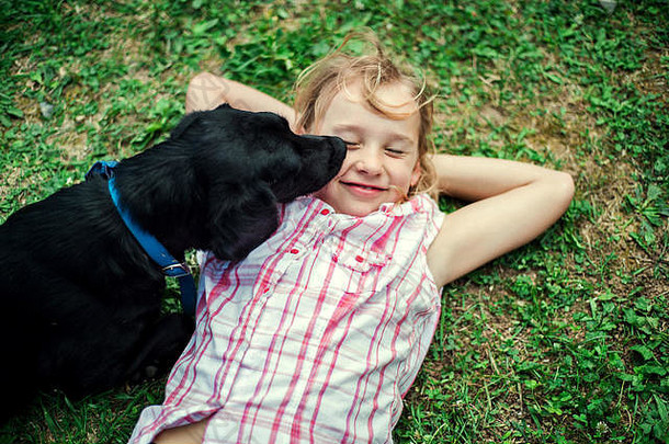 一个小女孩躺在草地上，一只黑狗在舔她的脸。