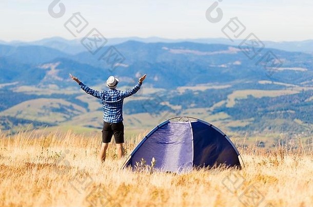 山顶上快乐旅行家的后视图，在户外露营帐篷附近，他举起双手享受自由和景色。旅游探险生活成功