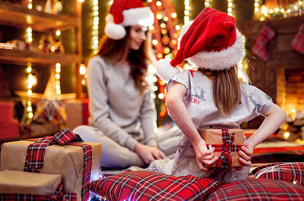 快乐的妈妈和她可爱的女儿戴着圣诞老人的帽子，穿着睡衣礼物。在圣诞节房间里用礼物爱家人。