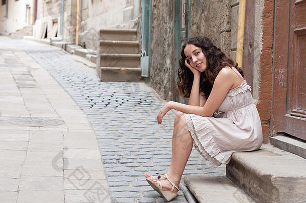在巴库老城区狭窄的街道上，穿着浪漫的比格裙的<strong>小卷发</strong>女孩坐在棕色木门前