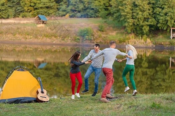 一群快乐的朋友带着吉他，在户外玩得很开心，在公园的湖边跳舞和跳跃，背景是美丽的天空。露营的乐趣。