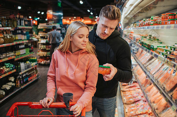 这对年轻夫妇在杂货店。男人手里拿着鱼子酱往下看。妇女站在旁边，拿着手推车。选择商店里的食物。