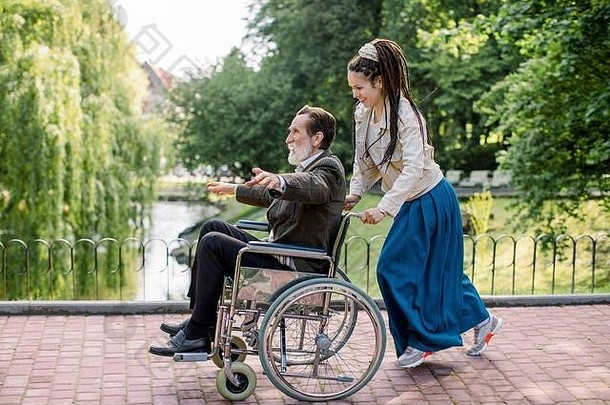 一位留着长发的漂亮时髦女郎推着轮椅和一位<strong>老人</strong>在湖边的城市公园里玩。残疾人