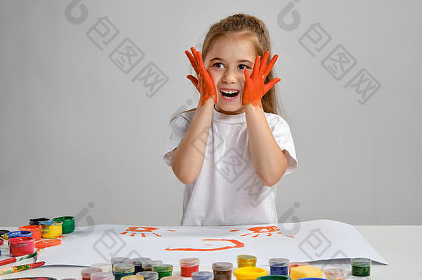 穿着白色t恤的小女孩坐在桌旁，桌上有惠特曼和五颜六色的颜料，画着她的脸颊。白色的隔离带。中等特写镜头。