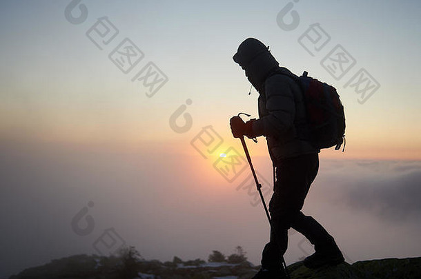 背着背包、拄着登山杖的徒步旅行者的剪影在洛基山顶徒步旅行，背景是雾蒙蒙的多云的蓝色天空，黎明时升起明亮的橙色太阳。