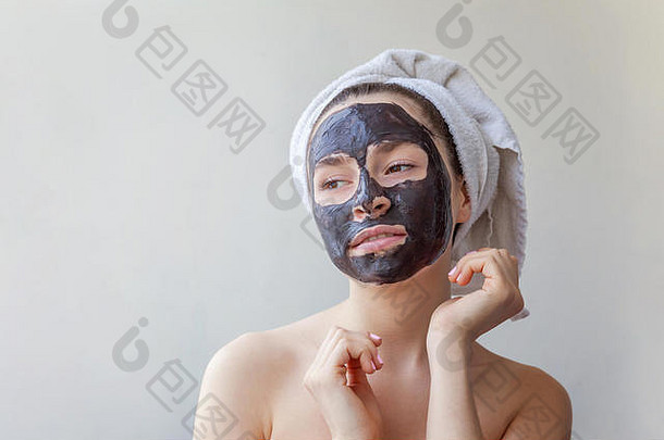 一位面带微笑的黑发女子的美丽肖像，头戴毛巾，在白色背景上脸上涂上黑色滋养面膜。护肤洁面spa放松化妆品概念