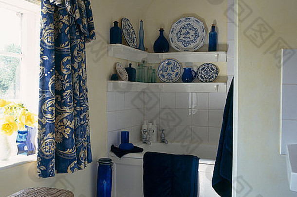 蓝色瓷器和玻璃器皿放在小浴室浴缸上方的架子上，带有蓝色和黄案的窗帘