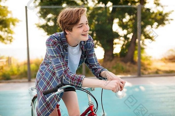 一个金发男孩穿着休闲衬衫，骑着红色自行车站在公园的篮球场上。年轻人高兴地一边看着一边站着