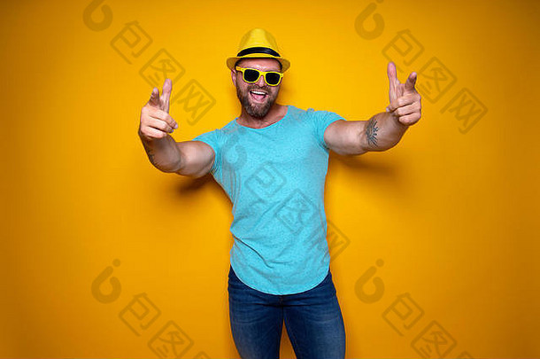 身穿蓝色衬衫、戴着墨镜、戴着黄色帽子的快乐男子站在黄色背景上，张开嘴微笑着，伸出手臂的手指指向前方，庆祝成功
