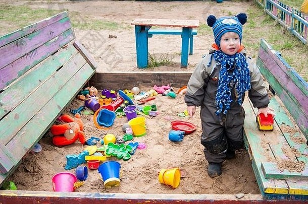 一个三岁的小男孩在一个凉爽的春天阳光明媚的日子里，在幼儿园的沙箱里玩耍，展示了他有多少玩具的各种情感——春天、凉爽、温暖的衣服
