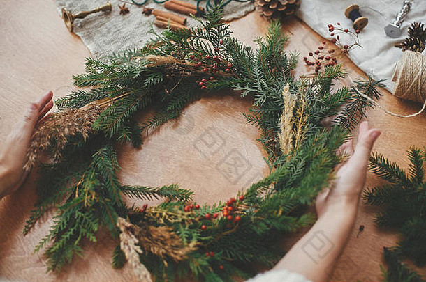 质朴的圣诞花环。手拿着圣诞花环，上面有冷杉枝、浆果、松果和线，剪刀放在乡村的木桌上。圣诞节