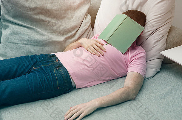 穿着牛仔裤和T恤的男人头戴着书睡在沙发上