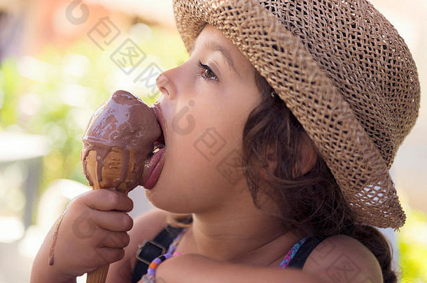 戴着草帽、穿着夏装的小女孩享受着夏日的酷暑，她吃着一个清爽的巧克力冰淇淋，奶油在手中融化