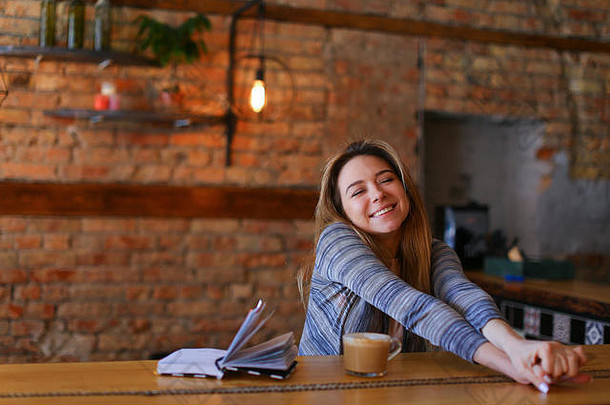 一个心满意足的女孩坐在桌子旁，端着一杯卡布奇诺和一本笔记本，坐在舒适的咖啡馆里，里面有着不同寻常的室内设计。