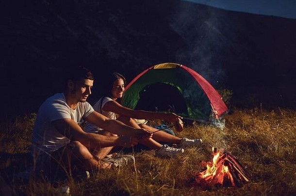 年轻的夫妇游客野营晚上烹饪面包篝火