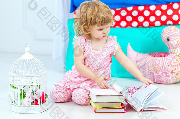 穿着粉色睡衣的可爱<strong>小卷发</strong>女孩坐在儿童卧室的地板上看书。