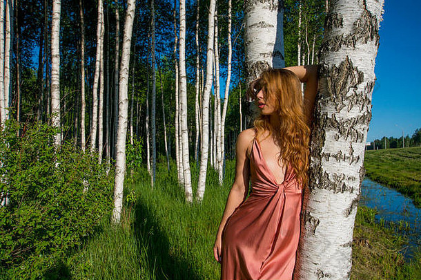 身穿珊瑚色长裙的年轻苗条美丽女孩在桦树林中摆姿势，手放在头上微笑着vogue stule