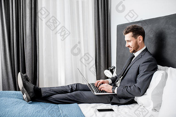 商务旅行期间，穿着西装的优雅商人在酒店房间的床上使用笔记本电脑