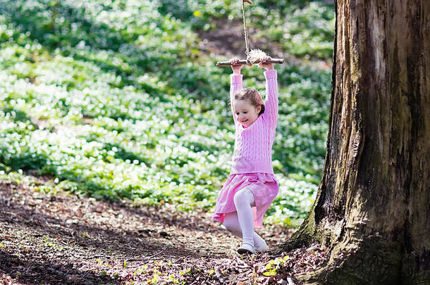 可爱的小女孩穿着粉红色的衣服在树绳上荡秋千，在盛开的春花花园里荡秋千。孩子们在后院的操场上荡秋千。孩子们玩游戏