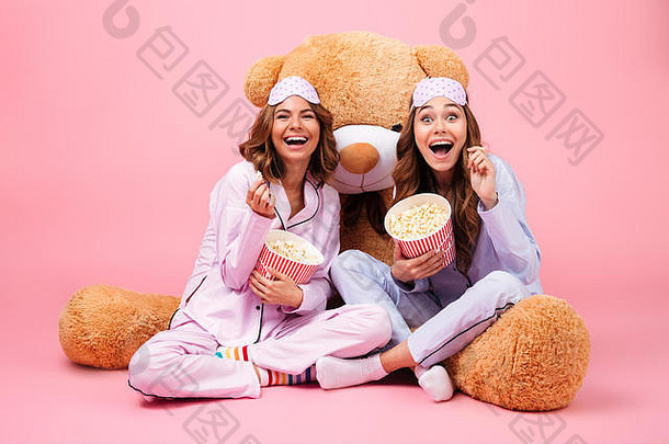 年轻的漂亮的女孩穿着睡衣笑坐着大泰迪熊吃爆米花孤立的粉红色的背景